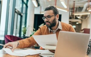 Man sifting through paper documents in front of his open laptop